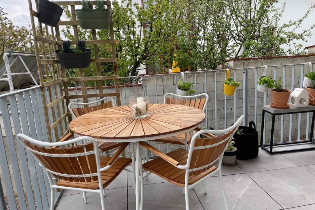 Balkon mit rundem Holztisch, vier Stühlen, hängenden Kräutertöpfen und vertikalem Pflanzregal.