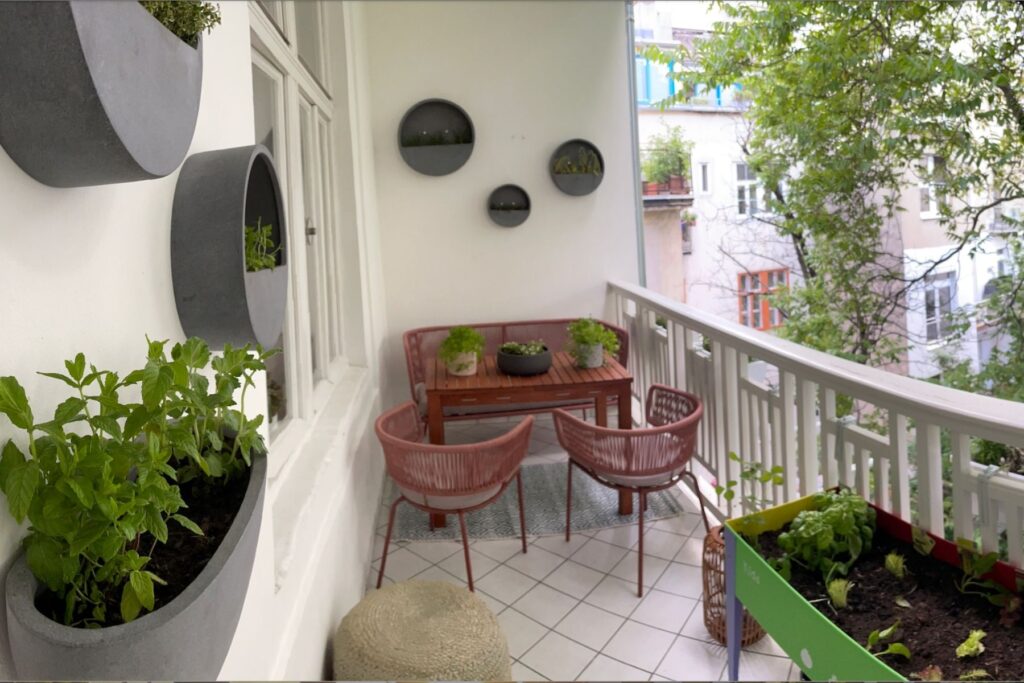 Schmaler Stadtbalkon mit Holztisch und zwei Sesseln, bepflanzten Wandgefäßen und grünem Hochbeet.
