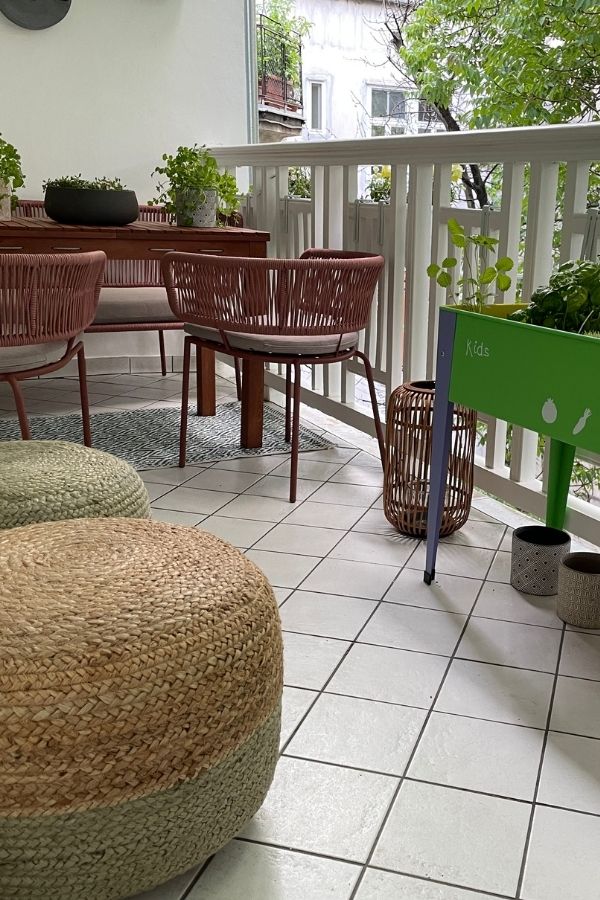 Gemütlicher Stadtbalkon mit Holztisch, zwei Sesseln, geflochtenen Poufs und grünem Hochbeet.