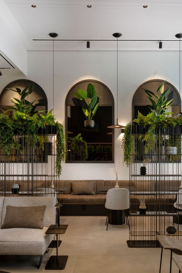 Moderner Loungebereich mit Sofas und begrünten Metallregalen vor Rundbogenfenstern.